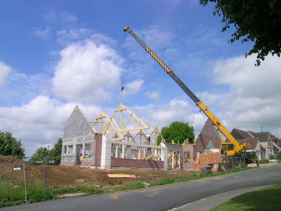 Chantier de construction avec une grue soulevant des fermes de toit en bois pour les poser sur une structure en briques sous un ciel nuageux.
