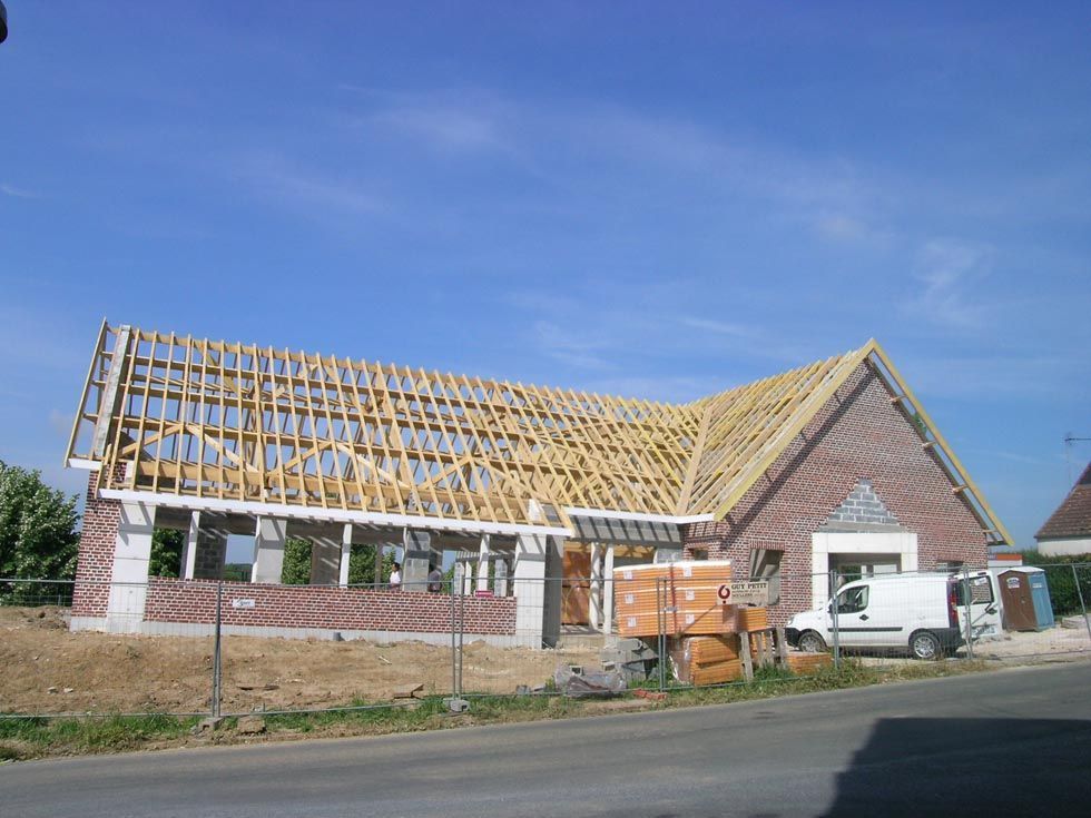Maison en construction, avec charpente de toit en bois apparente, murs en briques et une camionnette blanche garée devant.