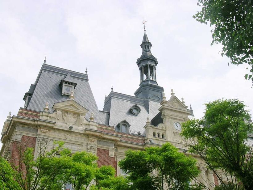 Bâtiment orné d'un haut clocher, d'une tour d'horloge et d'un toit gris foncé, partiellement encadré par des arbres verdoyants.