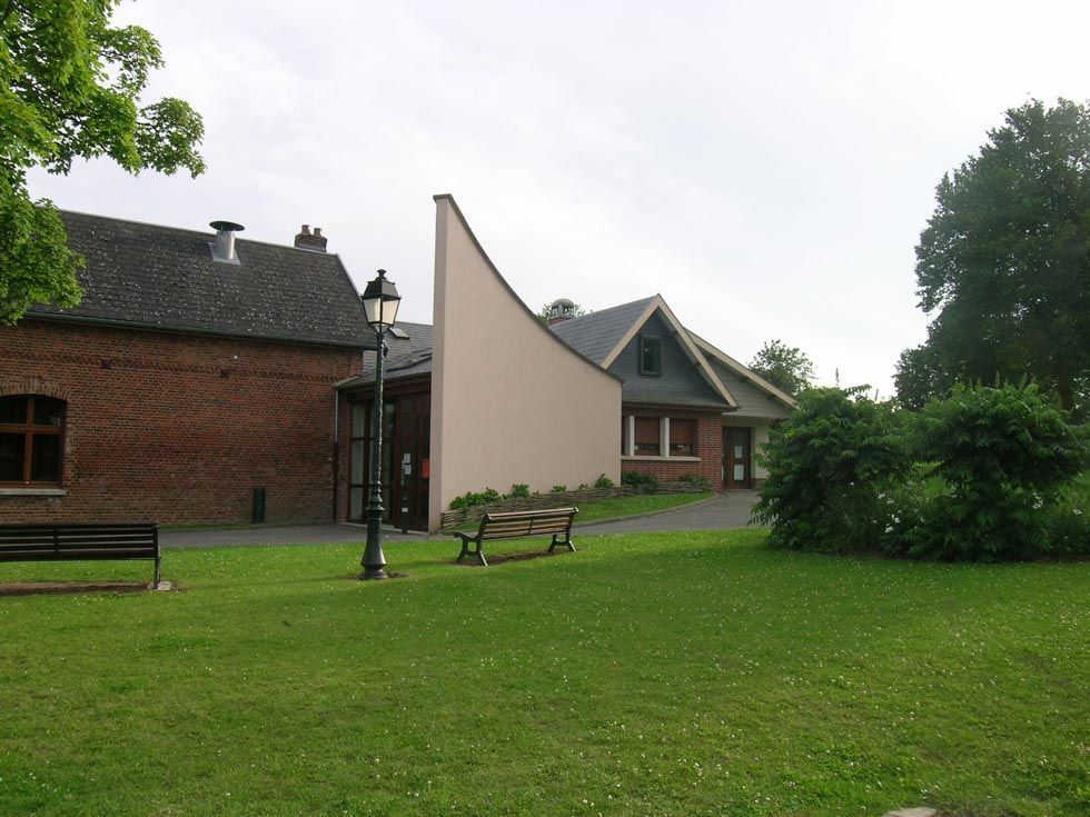 Des bâtiments en briques rouges et aux murs clairs se trouvent dans un parc verdoyant avec des bancs et un lampadaire.