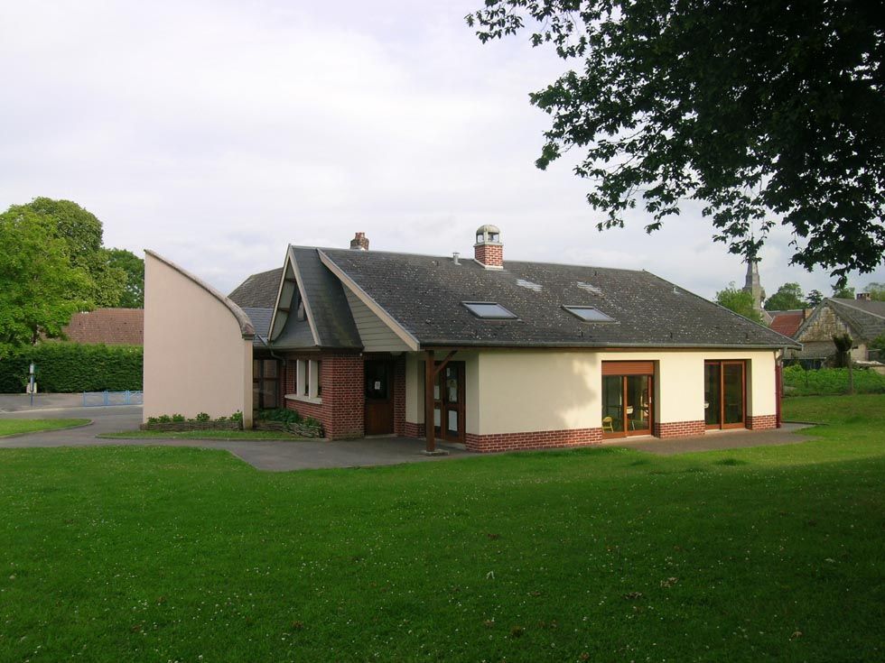 Bâtiment bas d'un seul étage, aux murs clairs, orné de briques rouges et d'un toit gris, dans un cadre verdoyant.