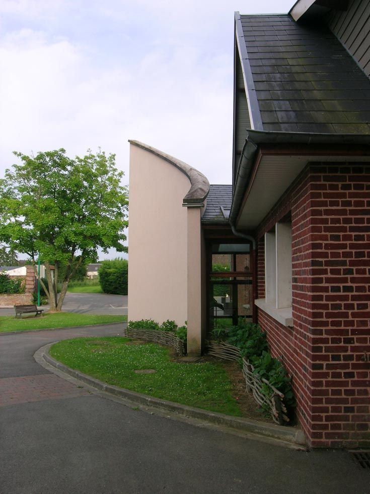Extérieur du bâtiment avec mur incurvé, briques rouges et toit sombre. Un arbre vert et de l'herbe bordent une zone pavée.