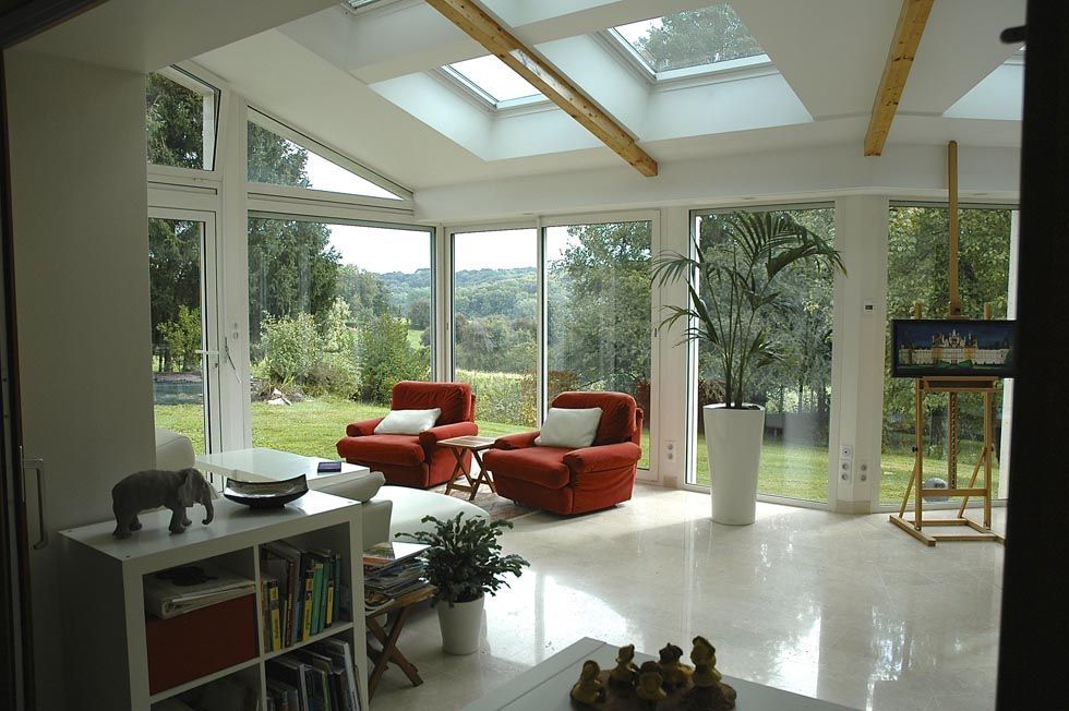 Véranda avec parois vitrées, puits de lumière, fauteuils rouges, donnant sur un jardin verdoyant et arboré.