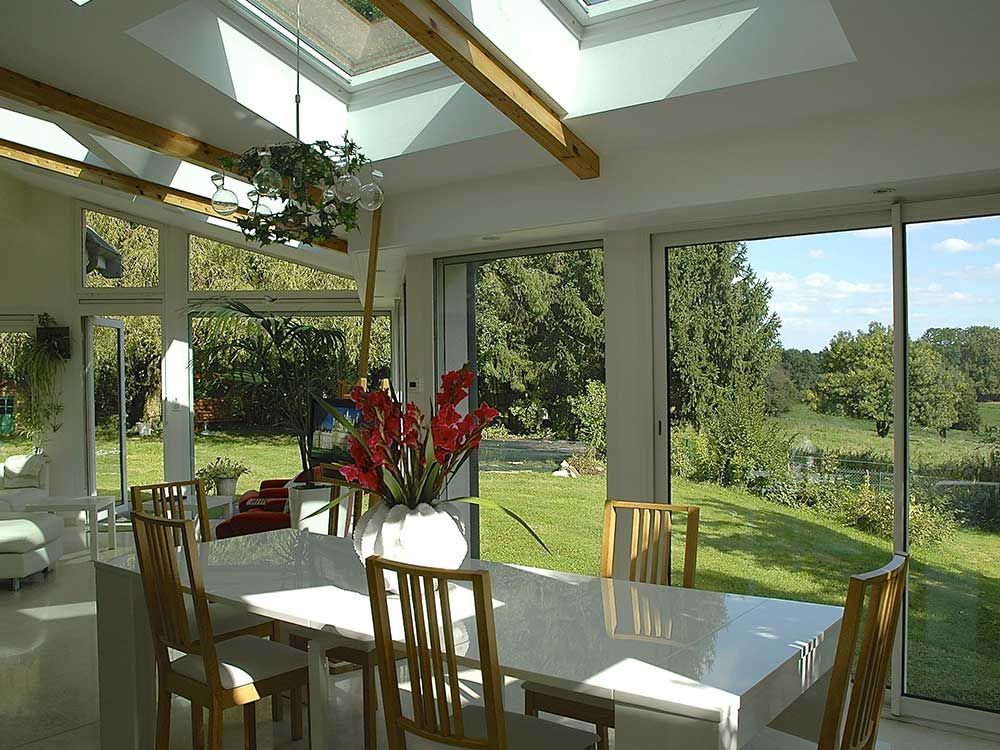 Salle à manger lumineuse avec une grande table blanche, des chaises et des portes coulissantes en verre donnant sur un jardin verdoyant et arboré.