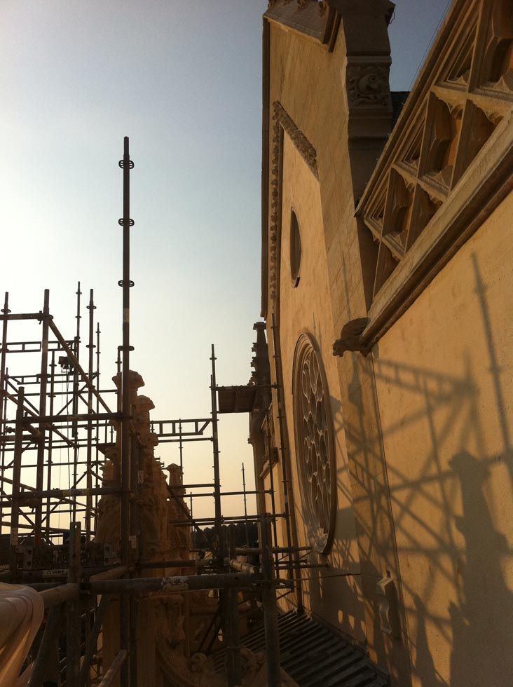 Extérieur d'un bâtiment en construction, échafaudages visibles. La lumière du soleil projette des ombres sur la façade en pierre ornée de détails décoratifs.