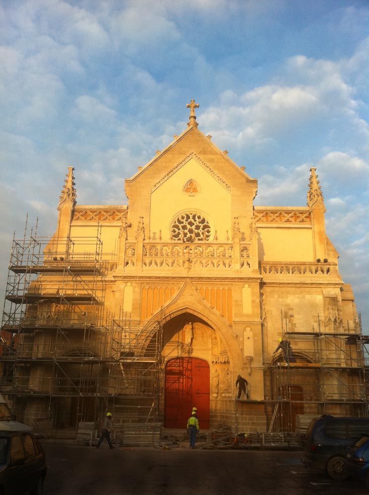 Façade d'église en construction ; échafaudages, portes rouges, ouvriers, ciel bleu.
