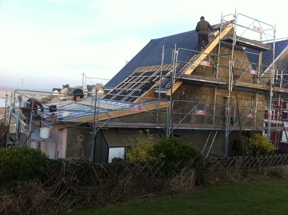 Des ouvriers du bâtiment installent de l'isolant sur le toit d'une maison. Des échafaudages entourent le bâtiment, dont une partie de la toiture est neuve.