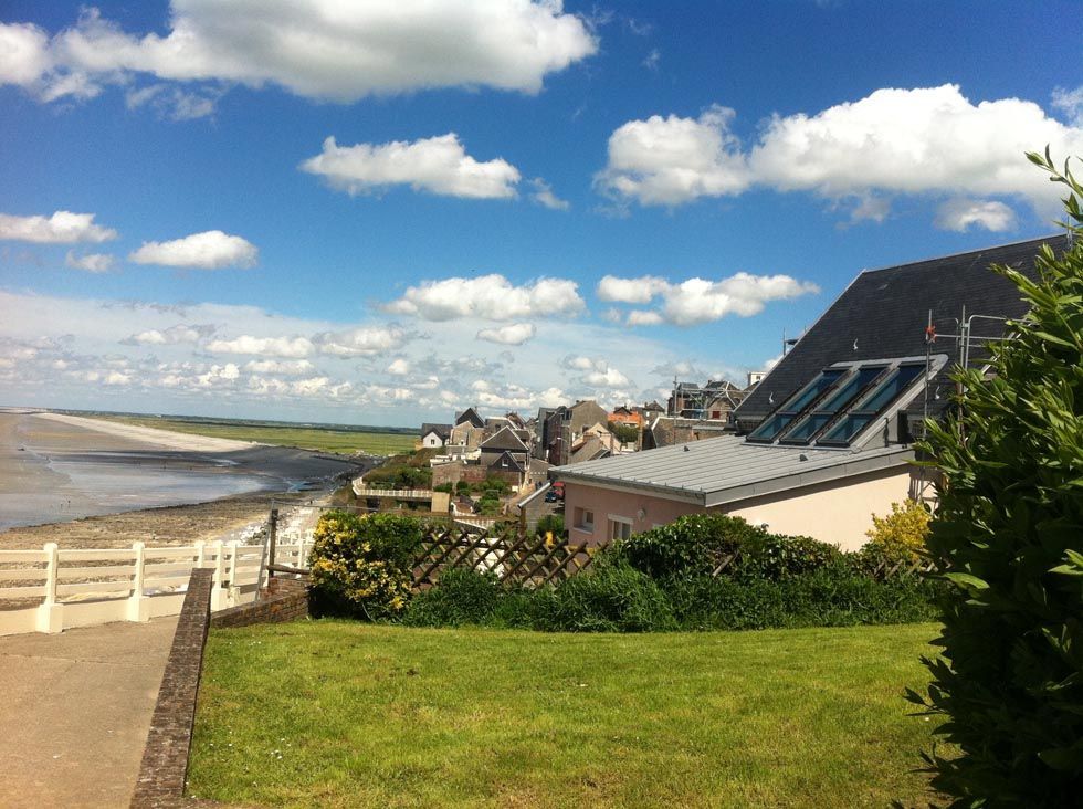 Colline herbeuse surplombant une plage et une ville sous un ciel bleu parsemé de nuages. Des bâtiments et la mer se dessinent au loin.