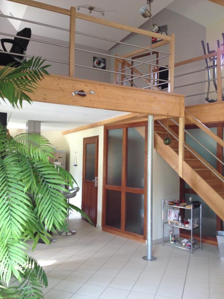 Intérieur avec mezzanine et escalier en bois, sol carrelé clair et grande fougère.