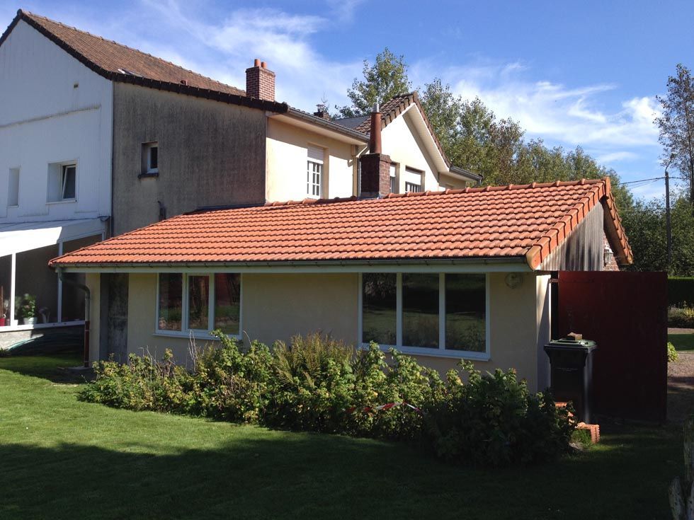 Bungalow beige clair au toit de tuiles rouges, jouxtant une maison plus grande de couleur beige. Pelouse et arbustes verts devant.