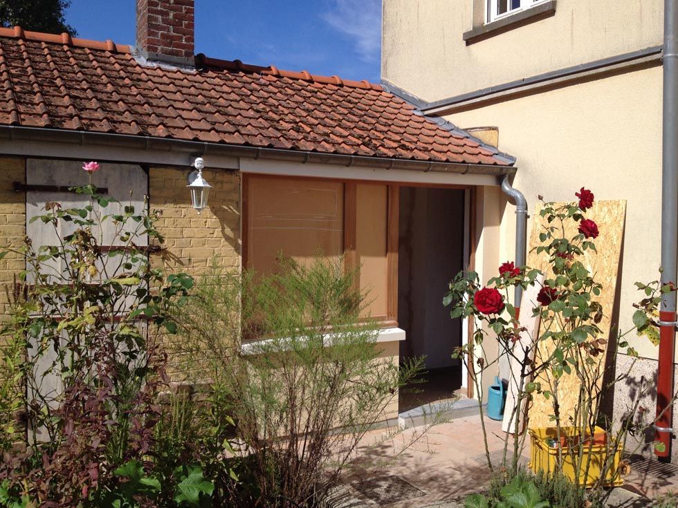 Petit bâtiment avec toit de tuiles, rosiers et porte partiellement condamnée.