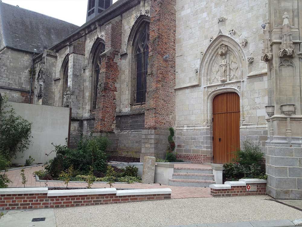 Vue latérale d'une ancienne église avec une porte en bois, une façade en briques et en pierres, et un jardin.