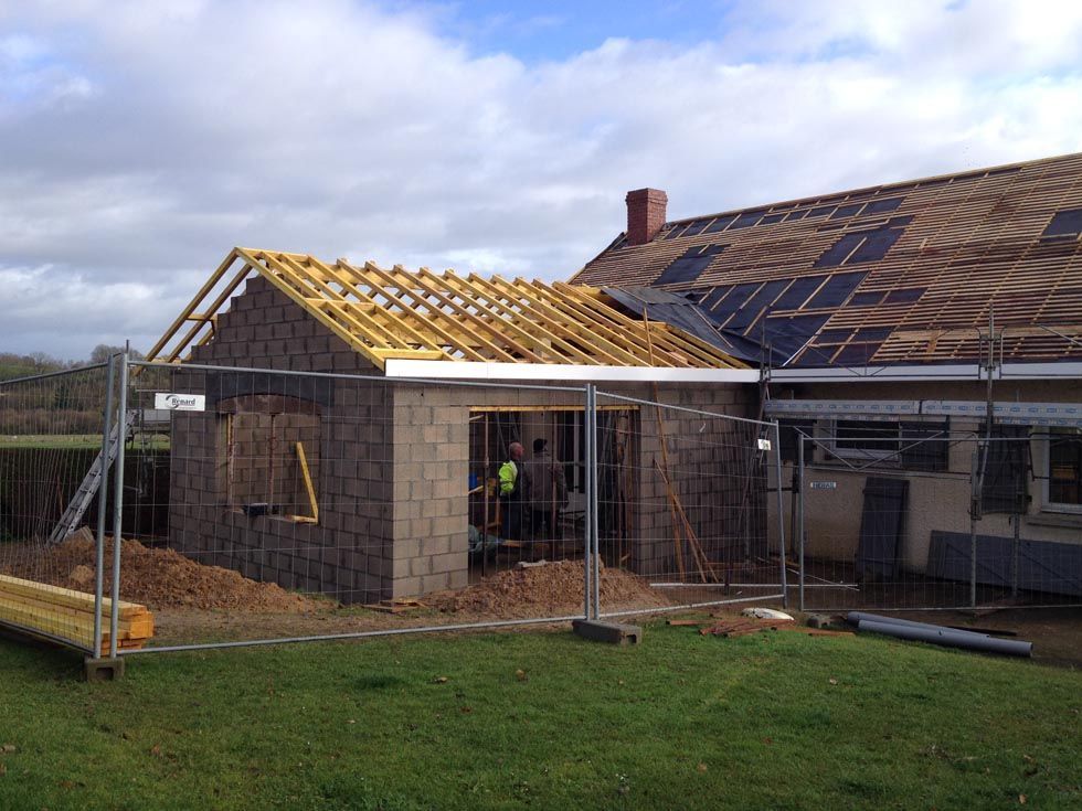 Chantier de construction avec extension en briques et toiture partiellement construites ; deux ouvriers visibles.