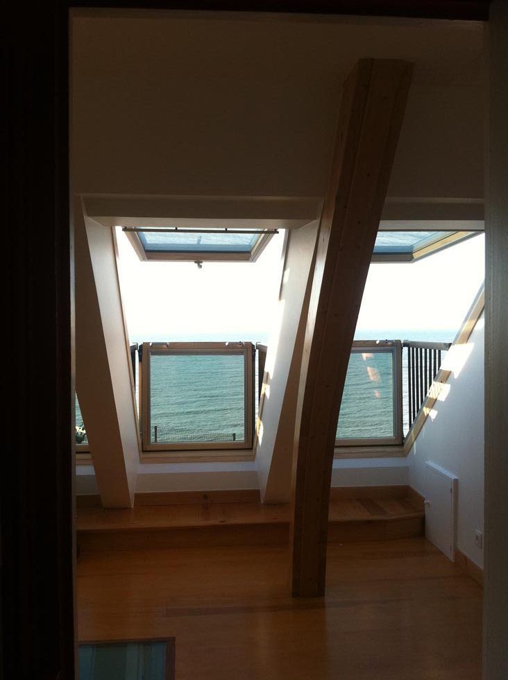 Vue intérieure d'une pièce avec des fenêtres en angle donnant sur la mer, une poutre de soutien en bois et un escalier.
