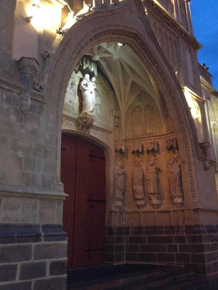 Entrée d'une église en pierre avec porte cintrée, porte rouge, statues et arche illuminée.