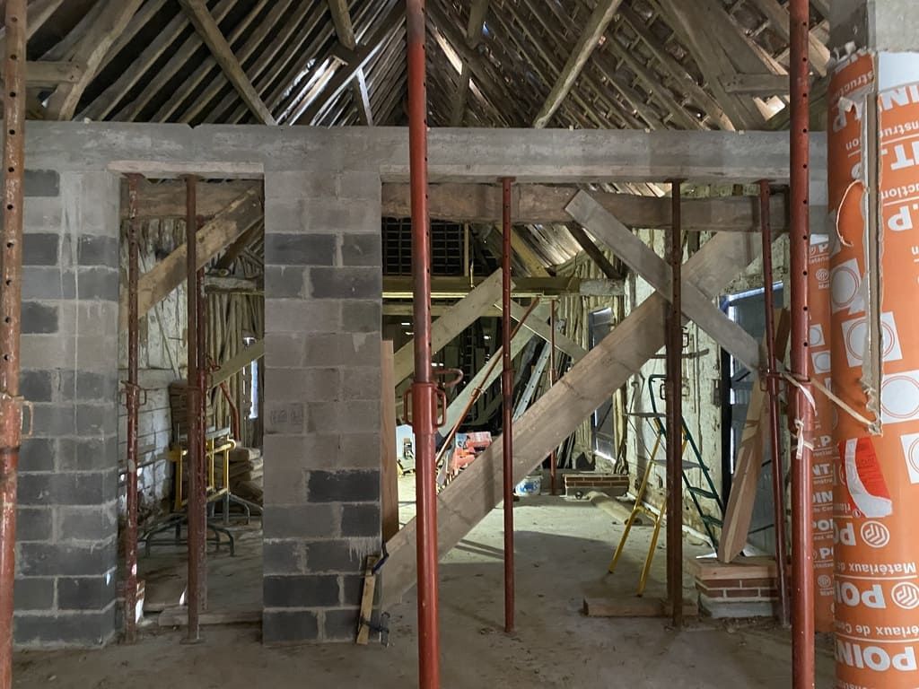 Intérieur d'un bâtiment en construction. Murs en parpaings percés d'ouvertures, soutenus par des échafaudages rouges. Poutres et toiture en bois visibles.