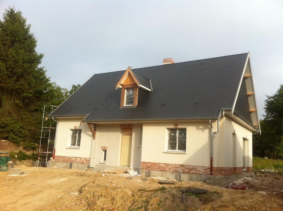 Maison en construction avec un toit foncé, des murs beiges et des détails en briques à la base.