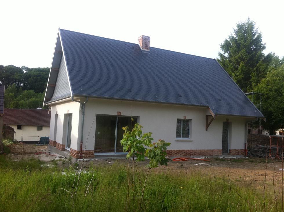 Maison blanche avec toit et cheminée sombres, en construction, jardin envahi par la végétation.