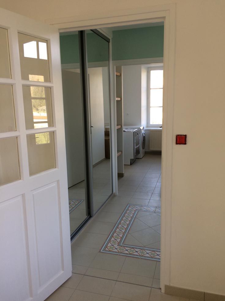 Une porte blanche ouverte donne sur une pièce dotée d'une porte coulissante à miroir, de murs clairs et d'un sol carrelé.