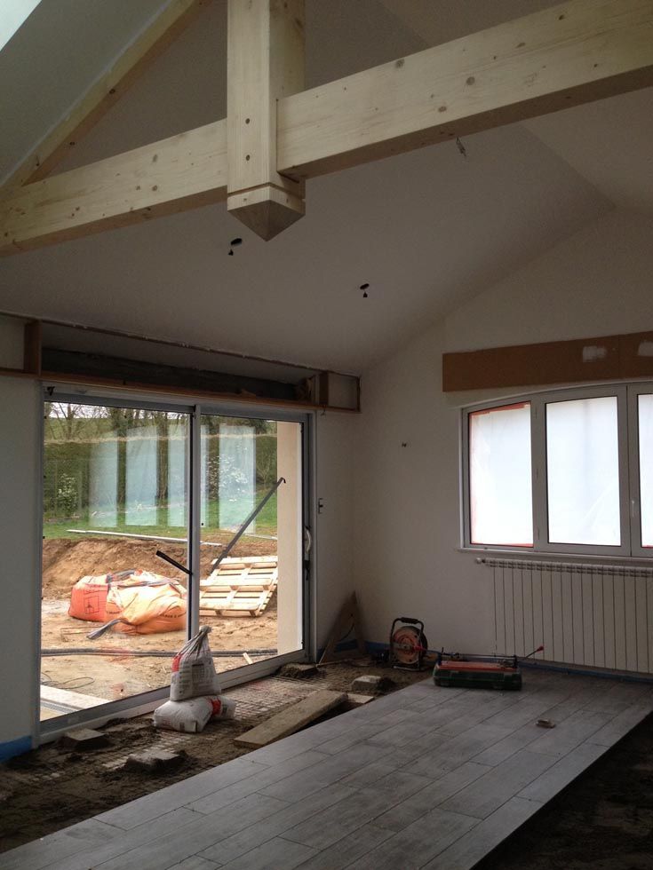Vue intérieure d'une pièce en construction avec poutres apparentes, grandes fenêtres et sol non fini.