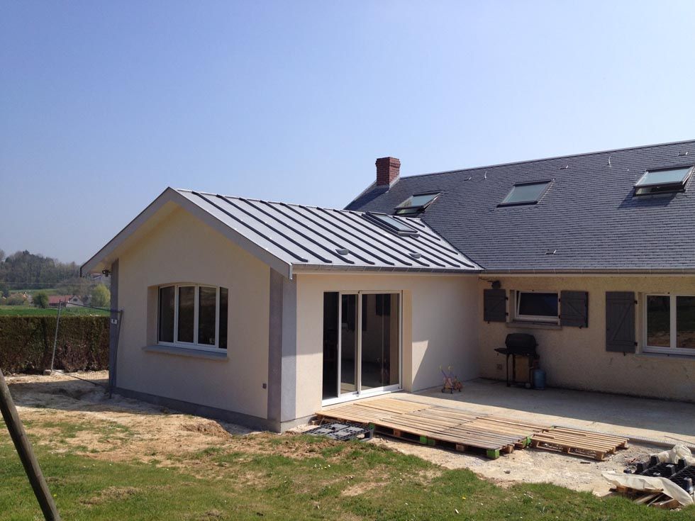 Maison beige avec une extension neuve, des portes coulissantes et un toit en métal, par une journée ensoleillée.