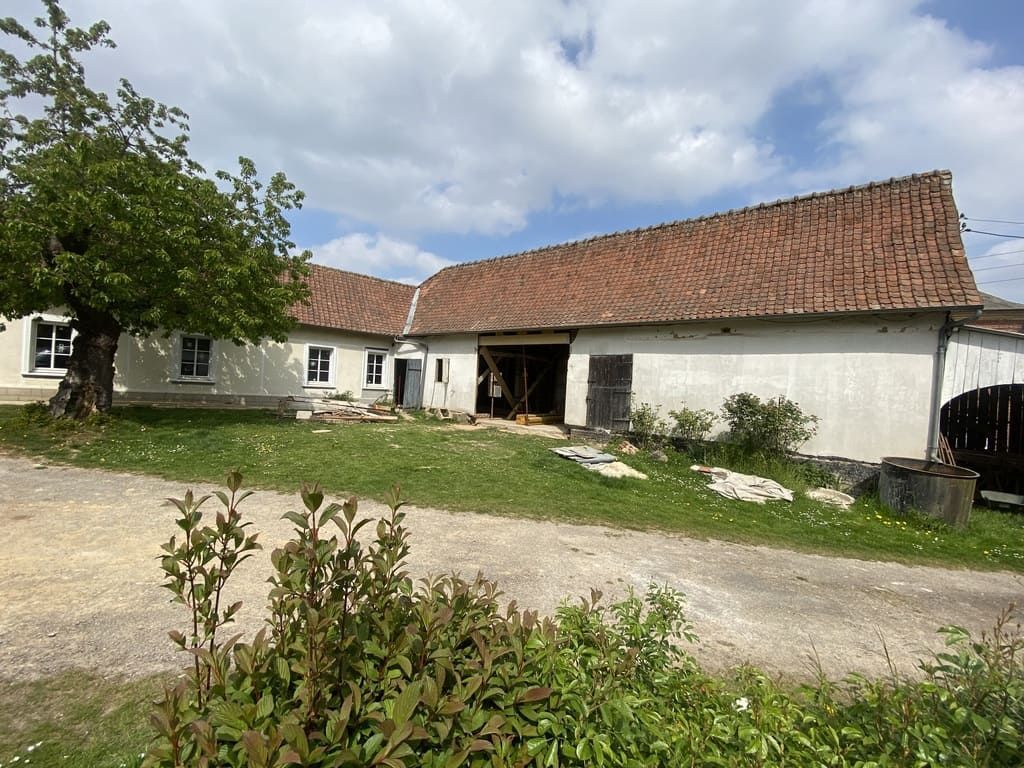 Un bâtiment rural blanc, au toit de tuiles rouges et à la grande porte de grange ouverte, sous un ciel nuageux.