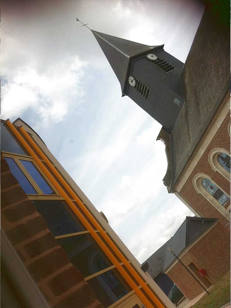 Église en briques avec un haut clocher, vue en angle. Bâtiment moderne aux accents jaunes à gauche. Ciel nuageux.