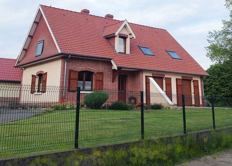 Maison au toit rouge, façade en briques beiges, volets marron, pelouse verte et clôture noire.