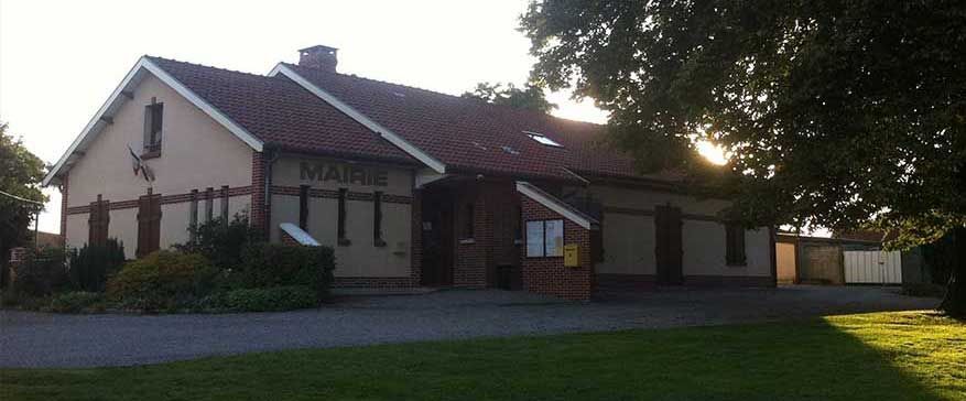 Un bâtiment beige avec un toit de tuiles rouges, une pelouse au premier plan et un grand arbre à droite.