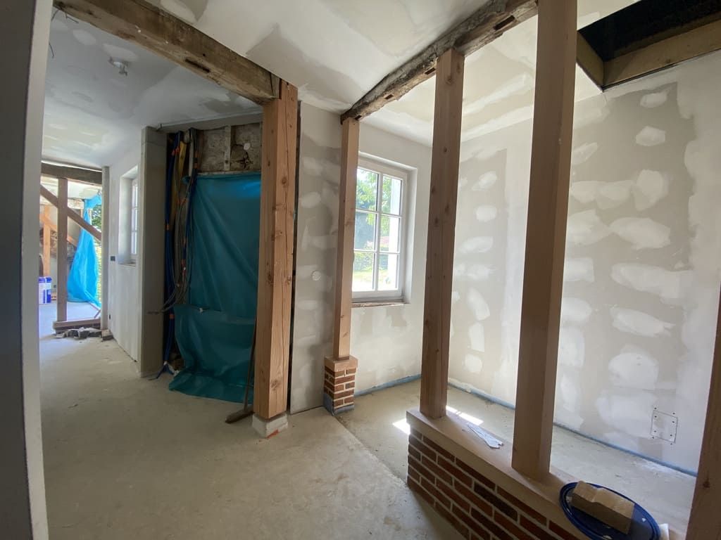 Chantier de construction intérieur ; poutres en bois encadrant les portes et les fenêtres, murs apparents, sol non fini.