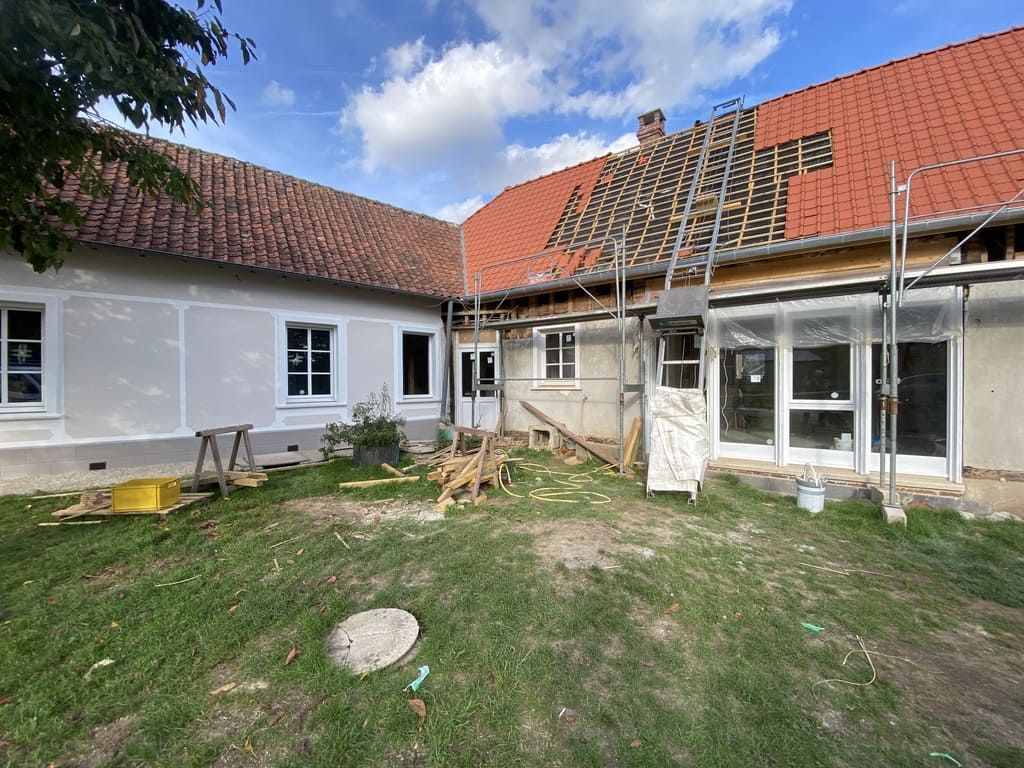 Extérieur d'un bâtiment en cours de réparation de toiture ; échafaudages, poutres apparentes et toit de tuiles rouges visibles.