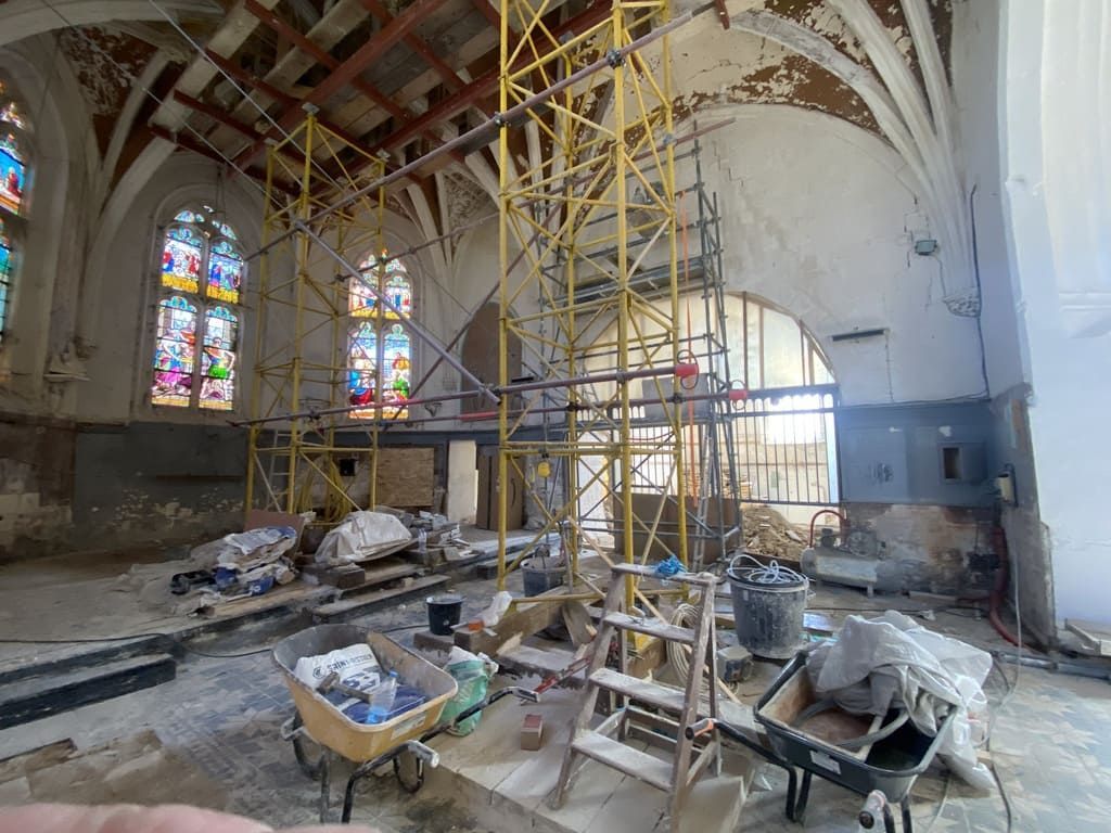 Intérieur d'un bâtiment en rénovation, avec des échafaudages, des vitraux et des débris de construction éparpillés un peu partout.