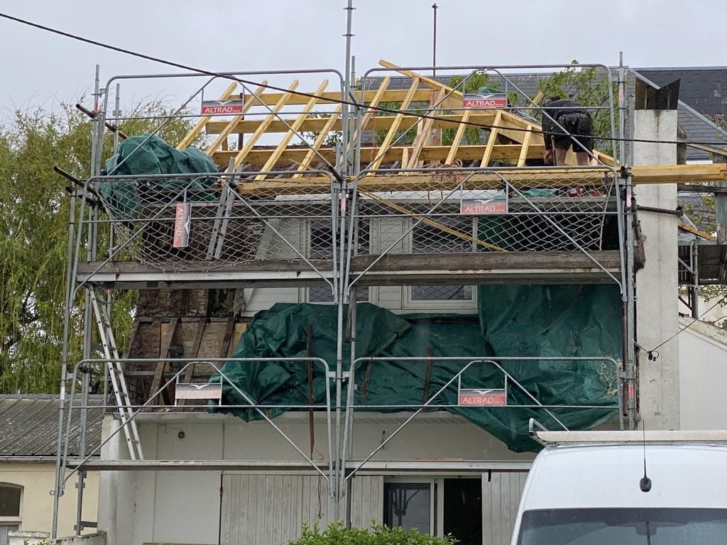 Des échafaudages entourent un bâtiment où la toiture est en cours de construction. Des bâches recouvrent certaines parties. Un ouvrier est visible.