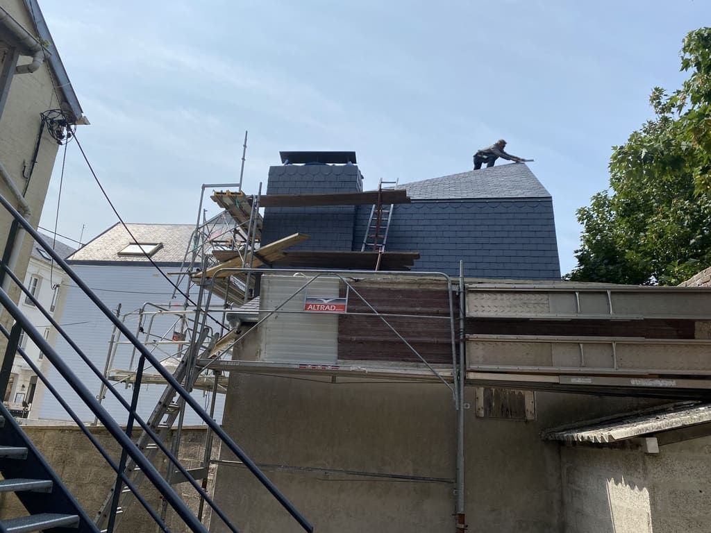 Couvreur sur un toit en ardoise, travaillant près de piles de matériaux. Échafaudage installé. Cadre extérieur ensoleillé.