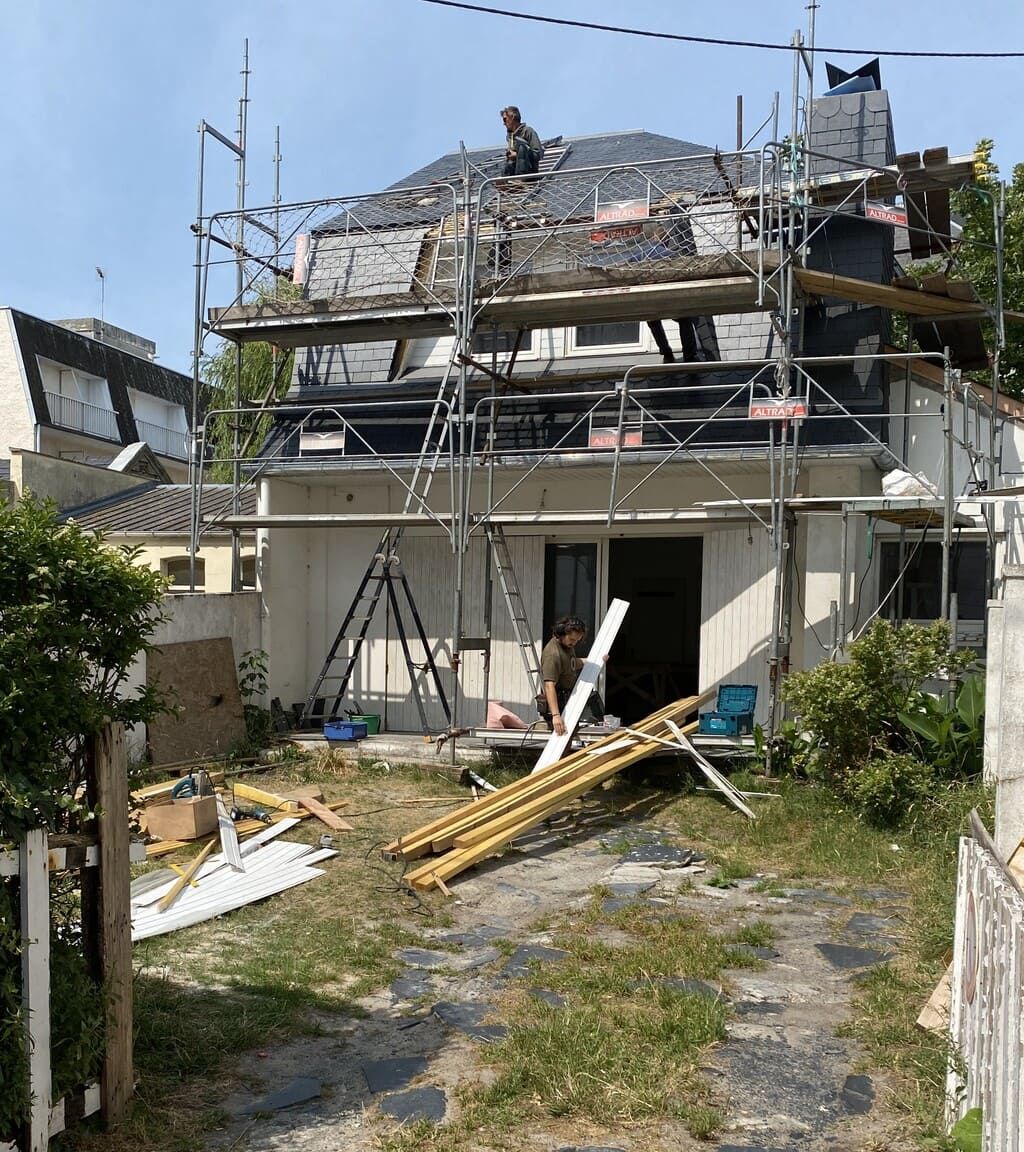 Maison en construction avec échafaudages. Ouvriers sur le toit, débris au sol.