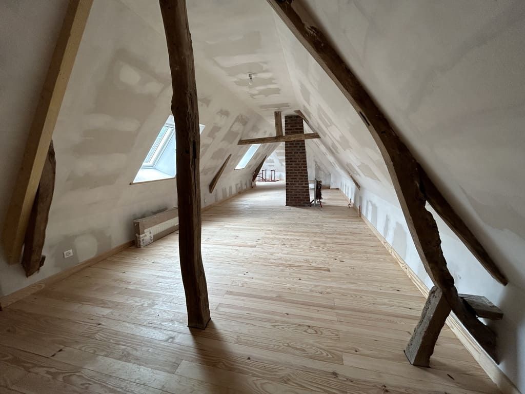 Combles aménagés avec poutres apparentes, cheminée et parquet clair.