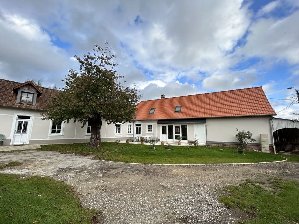 Maison de campagne avec toit de tuiles rouges, murs blancs, arbre dans la cour et ciel nuageux.