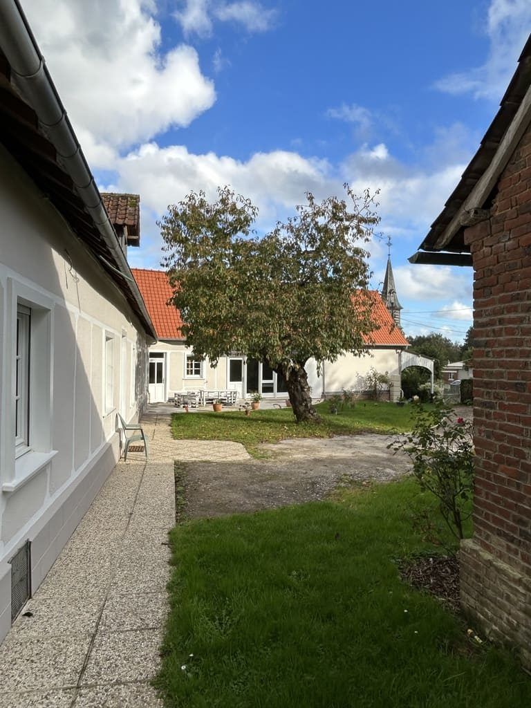 Cour intérieure entre les bâtiments, arbre au centre, clocher d'église visible en arrière-plan, ciel ensoleillé.