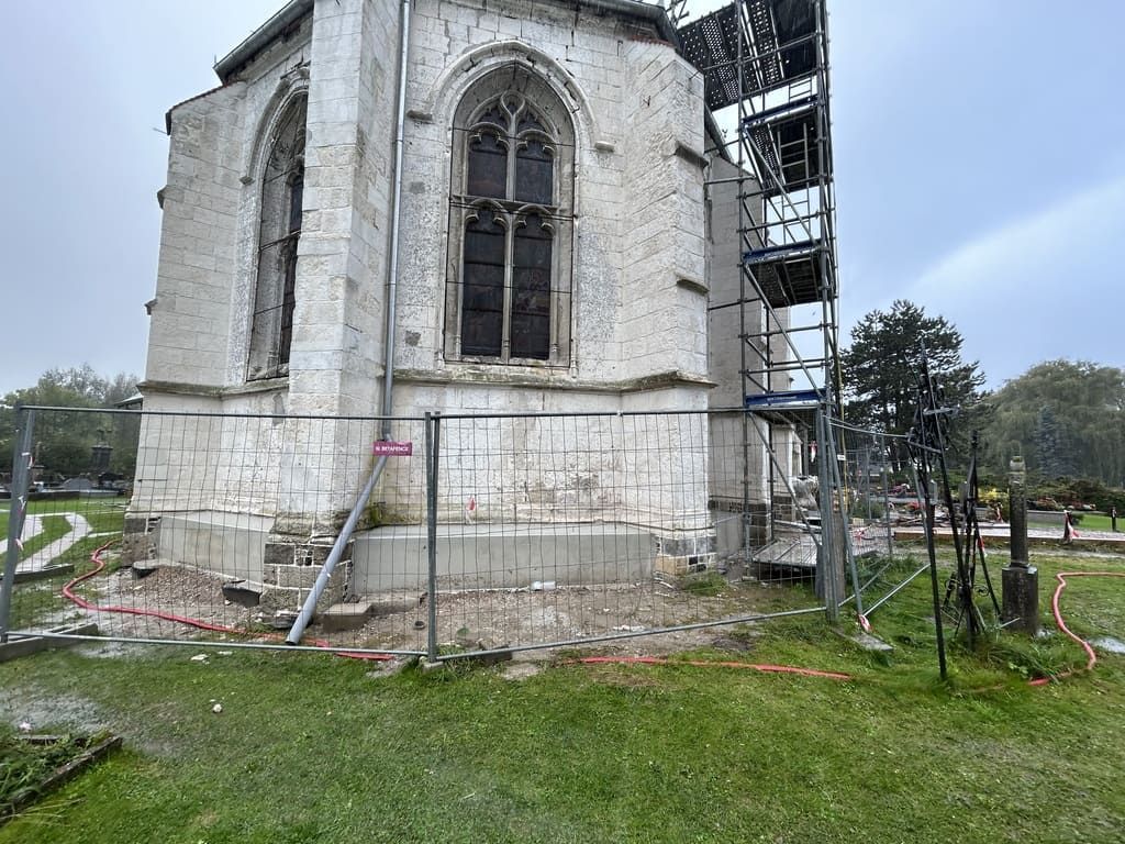 Vue extérieure d'une église endommagée en cours de réparation, avec échafaudages, barrière de sécurité et une zone herbeuse.