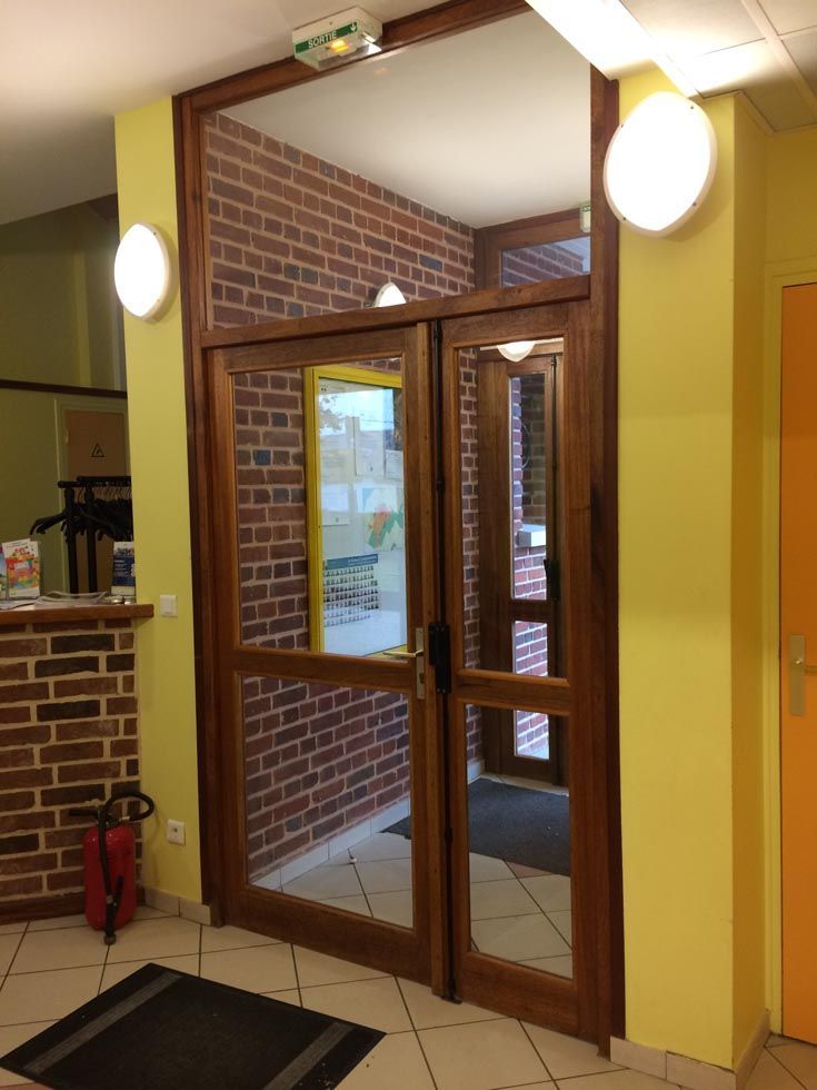 Double porte en bois avec panneaux de verre ; hall d'entrée avec mur de briques et peinture jaune.