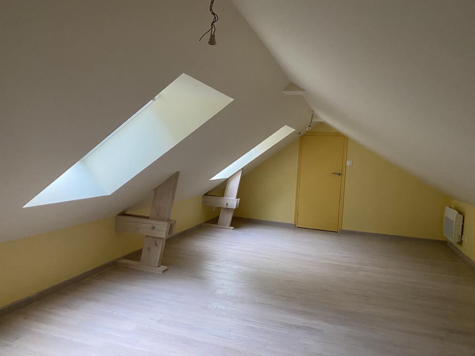 Pièce mansardée avec murs en pente, deux puits de lumière, une porte jaune, parquet et murs en bois clair.