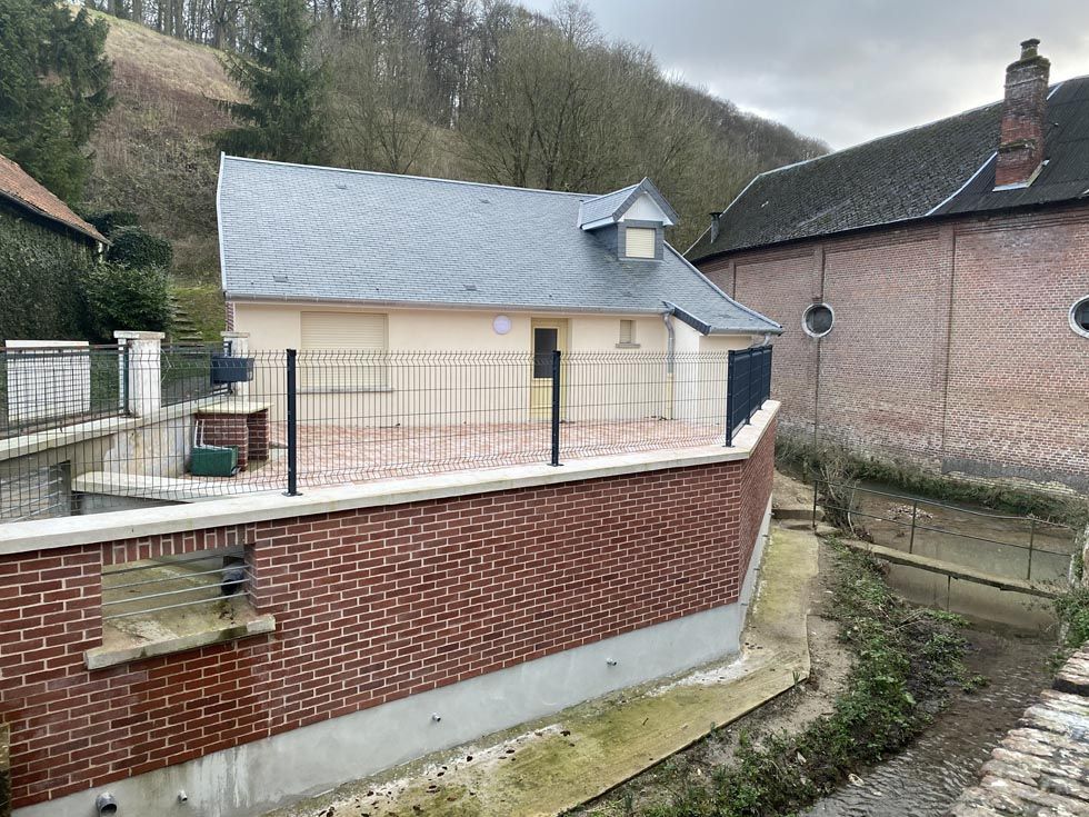 Petit bâtiment au toit gris, aux murs de briques et à la clôture noire. Un ruisseau coule devant.