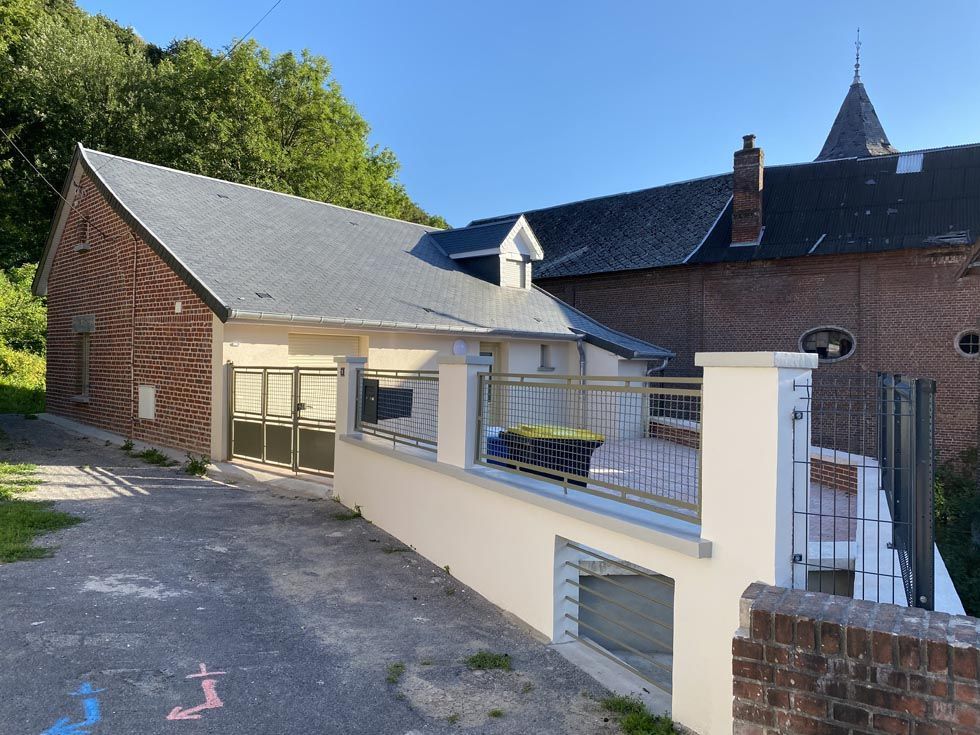 Bâtiment en briques au toit d'ardoise, petit bâtiment blanc, route et autres bâtiments, par une journée ensoleillée.