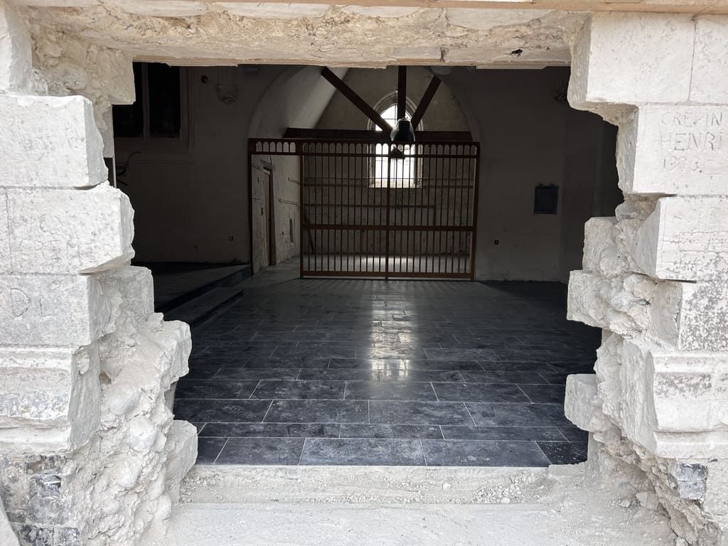 Une porte en pierre donne sur un intérieur avec un sol carrelé sombre, une grille et un ventilateur de plafond.