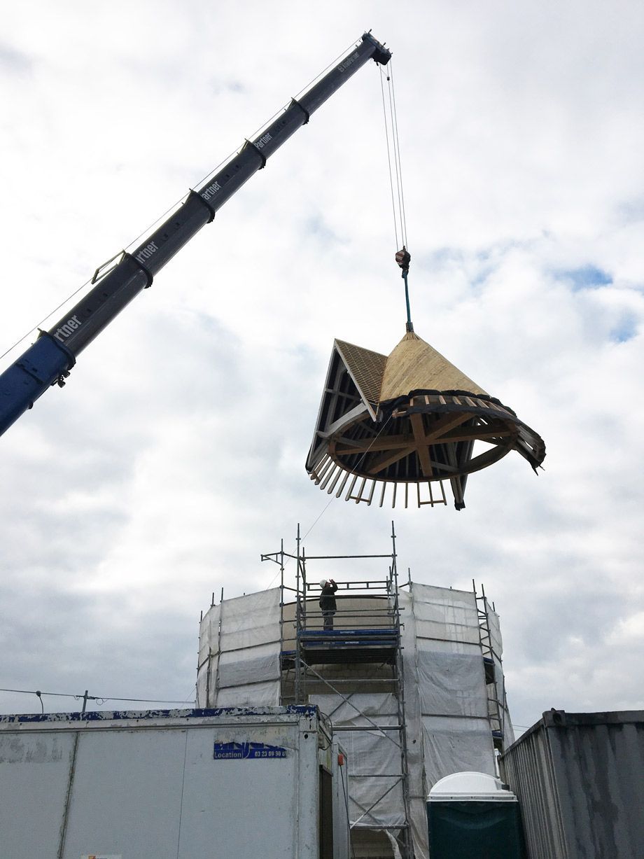 Une grue soulève une section de toiture en bois d'une tour pour l'installer au sommet d'une structure en pierre, entourée d'échafaudages.