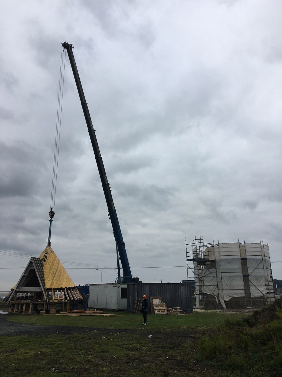 Grue soulevant une section de toiture sur un chantier, ciel nuageux. Une personne passe près d'un bâtiment en construction.