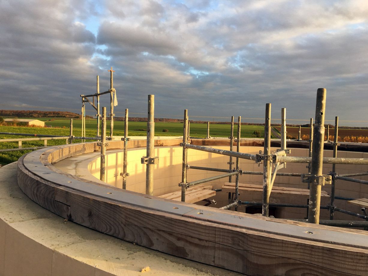 Structure circulaire en briques en construction, avec des échafaudages, se détachant sur un champ et un ciel nuageux.