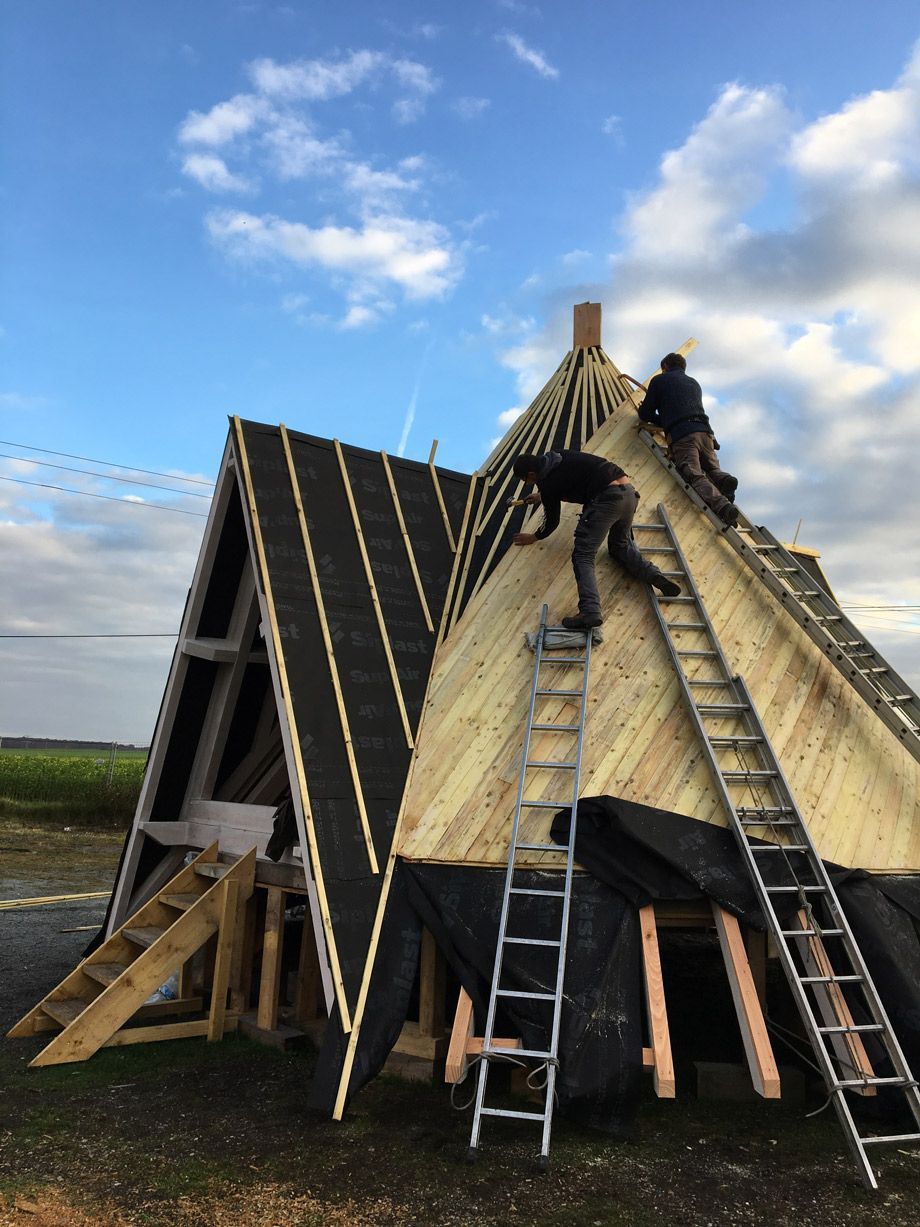 Deux ouvriers sur des échelles construisent une structure en bois en forme de A avec une toiture noire, sous un ciel nuageux.
