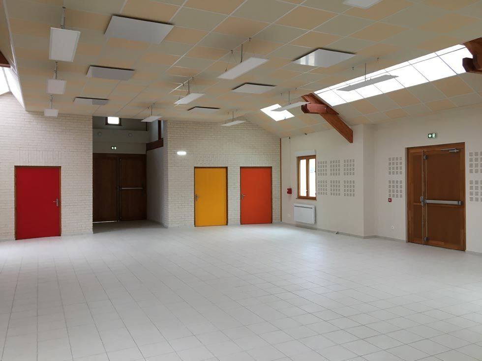 Salle communautaire vide avec des portes colorées, des murs blancs et des puits de lumière.