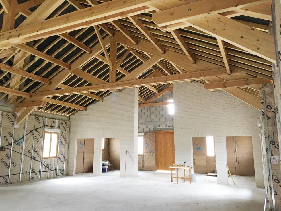 Vue intérieure d'un bâtiment en construction, montrant des poutres en bois apparentes, l'isolation et des murs partiellement finis.